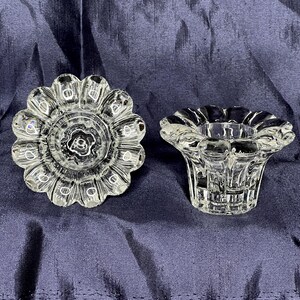 May include: Two clear glass candle holders with a decorative, scalloped design. The holder on the left has a circular base with a raised center. The holder on the right has a wider base with a flared top.