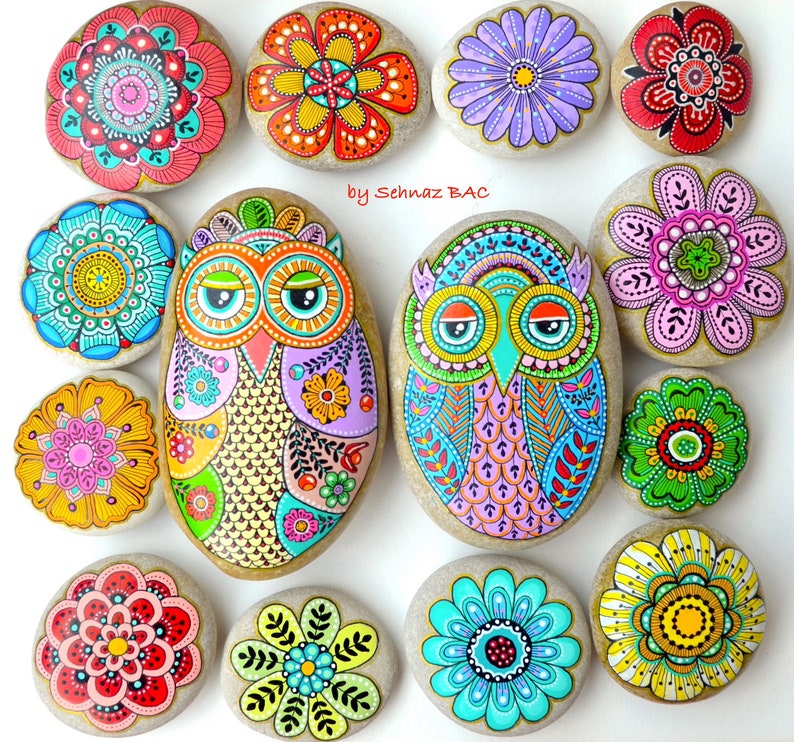 Puede incluir: Una colecci&oacute;n de piedras pintadas con dise&ntilde;os de b&uacute;hos, flores y mandalas. Las piedras est&aacute;n pintadas en colores brillantes, incluyendo rosa, azul, verde, amarillo y rojo. El texto "by Sehnaz BAC" est&aacute; escrito sobre el fondo blanco.