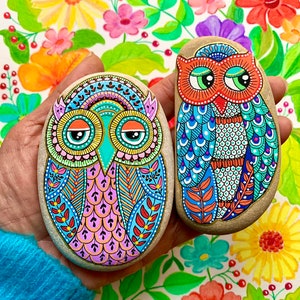 Puede incluir: Dos piedras pintadas con forma de b&uacute;ho con dise&ntilde;os coloridos e intrincados. Los b&uacute;hos tienen ojos grandes y redondos con pupilas negras y p&aacute;rpados naranjas. La piedra de la izquierda es rosa, azul y verde, mientras que la piedra de la derecha es azul, verde y naranja.