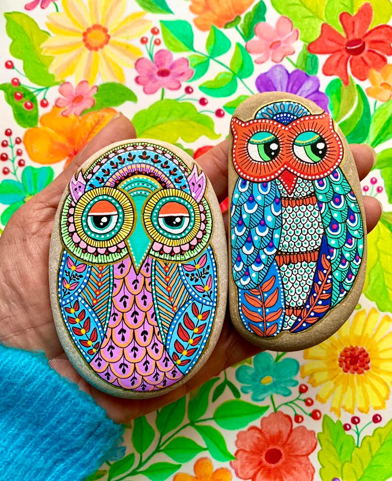 Puede incluir: Dos piedras pintadas con forma de b&uacute;ho con dise&ntilde;os coloridos e intrincados. Los b&uacute;hos tienen ojos grandes y redondos con pupilas negras y p&aacute;rpados naranjas. La piedra de la izquierda es rosa, azul y verde, mientras que la piedra de la derecha es azul, verde y naranja.