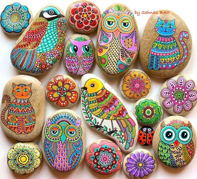 Puede incluir: Una colecci&oacute;n de 15 piedras pintadas a mano con varios dise&ntilde;os, incluyendo p&aacute;jaros, b&uacute;hos, flores y un gato. Las piedras est&aacute;n pintadas en colores brillantes y patrones intrincados. La firma del artista, "by Schnaz BAC", es visible en la esquina superior derecha de la imagen.