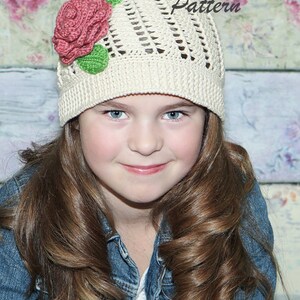 Puede incluir: Una persona joven lleva un gorro blanco de ganchillo con un adorno de flor rosa y verde. El gorro tiene un patrón de encaje y la persona tiene el pelo largo y castaño.