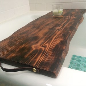 May include: A dark brown wooden bath tray with a leather handle and brass accents. The tray is sitting on a white bathtub.