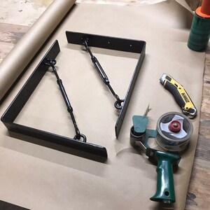 May include: Two black metal shelf brackets with chain supports, a green tape dispenser, a yellow utility knife, and a roll of brown paper on a wooden surface.