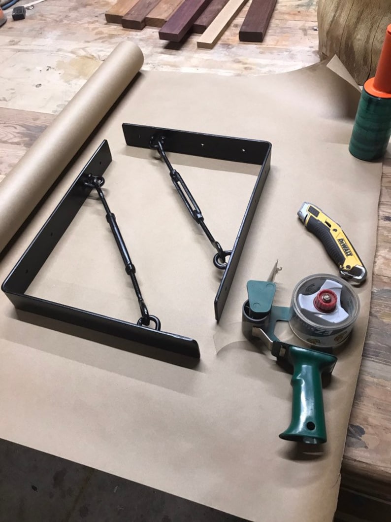 May include: Two black metal shelf brackets with chain supports, a green tape dispenser, a yellow utility knife, and a roll of brown paper on a wooden surface.