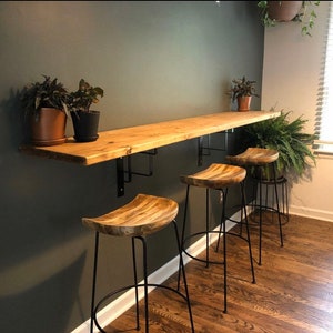 May include: A wooden floating shelf with black metal brackets mounted to a dark green wall. Three black metal bar stools with wooden seats are positioned in front of the shelf.