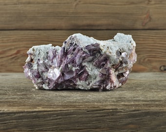 May include: A large, rough, purple and white crystal cluster on a wooden surface. The crystal has a natural, raw appearance.