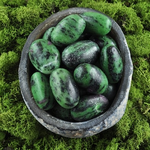 May include: A bowl filled with green and black zebra jasper tumbled stones. The stones are smooth and polished, and they are arranged in a random pattern. The bowl is made of dark grey ceramic and has a rough texture.