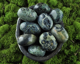 May include: A collection of green and black ocean jasper tumbled stones in a small wooden bowl. The stones are smooth and polished, with a variety of patterns and swirls.