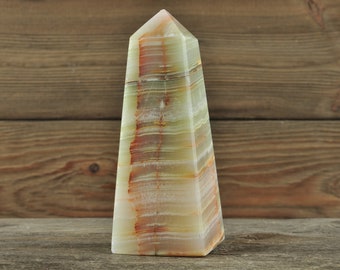 May include: A polished onyx obelisk with a pyramidal top. The stone features bands of green, cream, and reddish-brown hues. The obelisk is displayed on a wooden surface against a wood-panelled backdrop.
