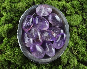 May include: A bowl filled with amethyst tumbled stones, the purple stones are smooth and polished, the bowl is sitting on a bed of green moss.