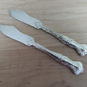 May include: Two silver butter knives with ornate handles. The knives feature a decorative design of grapes and leaves.
