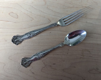 May include: A silver fork and spoon with ornate floral designs. The utensils are laid out on a wooden surface.