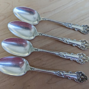 May include: Four silver teaspoons with ornate handles. The spoons have a polished finish and are arranged in a row on a wooden surface.