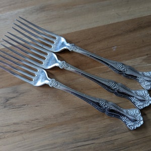 Puede incluir: Tres tenedores plateados con mangos ornamentados, con un diseño de racimo de uvas. Las púas son largas y delgadas. Los tenedores están dispuestos diagonalmente sobre una superficie de madera, mostrando sus elegantes detalles.