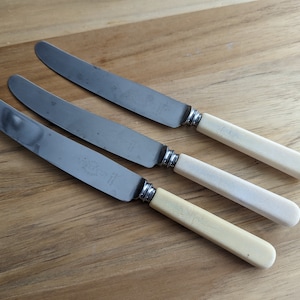 May include: Three silver butter knives with ivory handles. The knives have a slightly curved blade and a rounded tip.