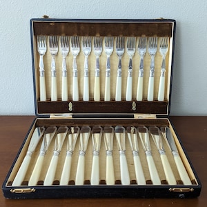 Puede incluir: Una caja de cuero negro con forro de terciopelo contiene un juego de 24 tenedores y cuchillos de plata con mangos de marfil.