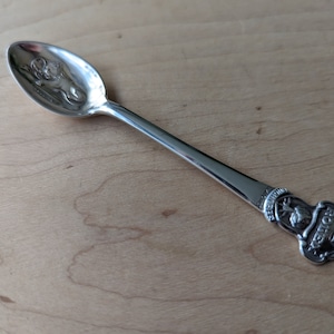 Puede incluir: Una cucharita de plata con un diseño grabado de un león en el cuenco y un diseño detallado de un león y un árbol en el mango. El mango está inscrito con las palabras "The Lion's Share".