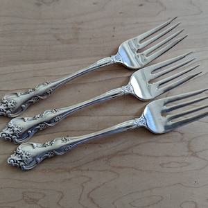 May include: Four silver forks with ornate handles. The forks are arranged in a row on a wooden surface.