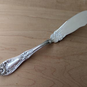 May include: A silver butter knife with a floral design on the handle and a scalloped blade. The knife is made of a shiny metal and rests on a wooden surface. The handle features detailed floral patterns.