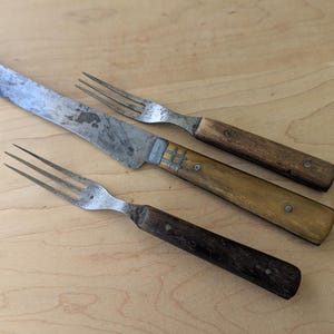 May include: An antique cutlery set featuring a large knife and two forks. The knife has a long, worn blade and a light-colored handle with metal accents. The forks have three tines and wooden handles, all set against a light wood surface.