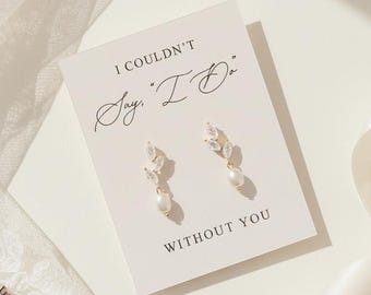 May include: Gold-coloured earrings with a leaf design and a pearl drop, presented on a card with the text "I Couldn't Say 'I Do' Without You." A rose gold ring and white ribbon are also visible.