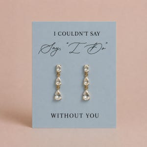 May include: A pair of gold-tone drop earrings featuring three clear, faceted stones in oval and teardrop shapes. The earrings are displayed on a light blue card with the black printed text: I COULDN'T SAY "Say, 'I Do'" WITHOUT YOU.