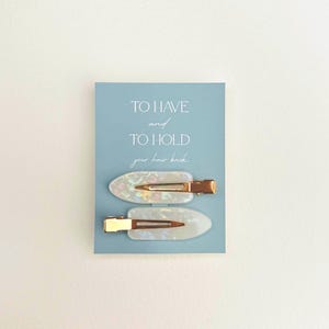 May include: Two pearlescent hair clips with gold-colored metal clasps are displayed on a light blue card. The card has the text "TO HAVE and TO HOLD your hair back" in a delicate font. The clips are oval-shaped.