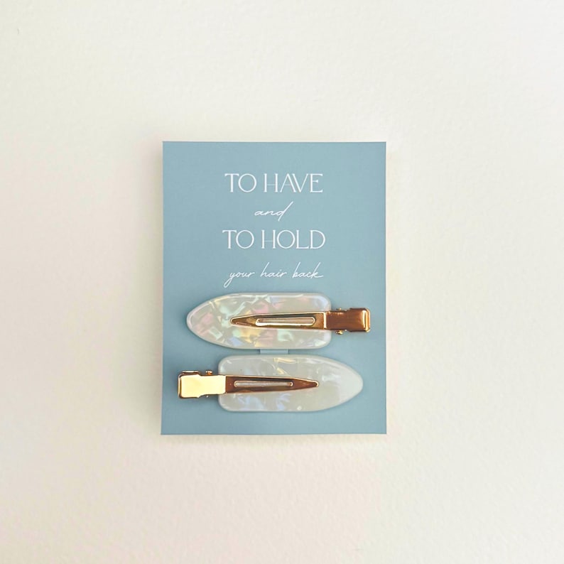 May include: Two pearlescent hair clips with gold-colored metal clasps are displayed on a light blue card. The card has the text "TO HAVE and TO HOLD your hair back" in a delicate font. The clips are oval-shaped.