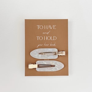 May include: Two white and pearlescent hair clips with gold-colored metal clasps are displayed on a brown card. The card has the text "TO HAVE and TO HOLD your hair back" in a handwritten style font.
