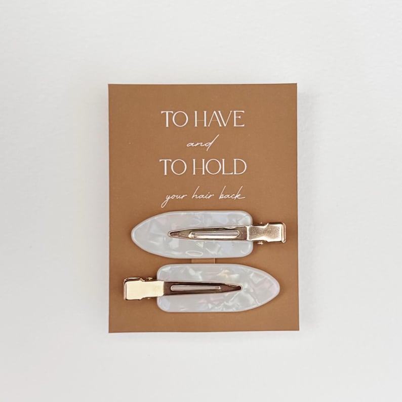 May include: Two white and pearlescent hair clips with gold-colored metal clasps are displayed on a brown card. The card has the text "TO HAVE and TO HOLD your hair back" in a handwritten style font.