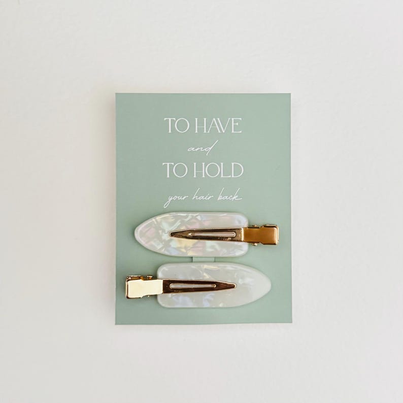 May include: Two pearlescent white hair clips with gold-colored metal clasps are displayed on a light green card. The card has the text "TO HAVE and TO HOLD your hair back" in a mix of fonts. The clips are oval-shaped.