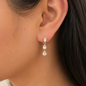 May include: A gold drop earring featuring three clear, faceted, pear-shaped stones arranged vertically. The stones are set in gold metal prongs and connected by small gold links, creating a delicate, dangling design that hangs from the earlobe.