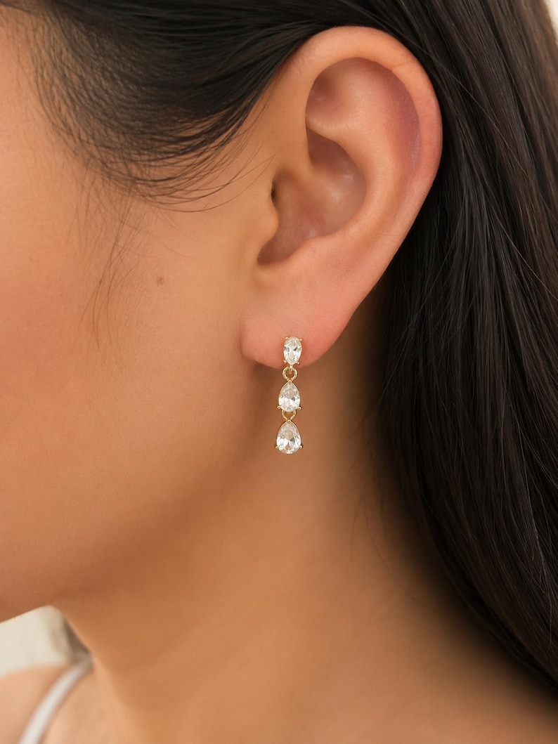 May include: A gold drop earring featuring three clear, faceted, pear-shaped stones arranged vertically. The stones are set in gold metal prongs and connected by small gold links, creating a delicate, dangling design that hangs from the earlobe.