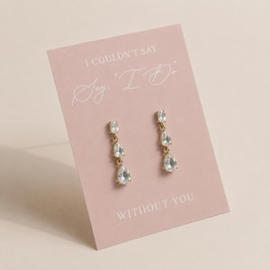 May include: A pair of gold-tone drop earrings featuring three clear, pear-shaped crystals on a light pink display card. The card displays the text I COULDN'T SAY "I DO" WITHOUT YOU in white lettering, presenting the jewelry as a gift for a wedding.