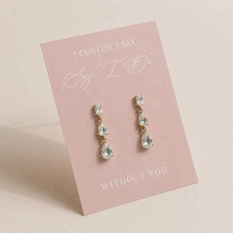 May include: A pair of gold-tone drop earrings featuring three clear, pear-shaped crystals on a light pink display card. The card displays the text I COULDN'T SAY "I DO" WITHOUT YOU in white lettering, presenting the jewelry as a gift for a wedding.