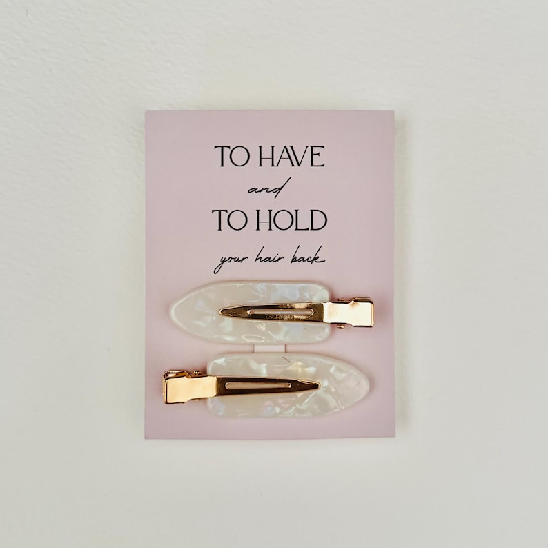 May include: Two white and gold hair clips on a pink card. The clips have an oval shape with a pearlescent finish and gold-colored metal clasps. The card has the text "TO HAVE and TO HOLD your hair back."
