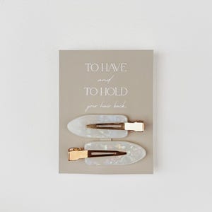 May include: Two white and gold hair clips on a light gray card. The clips have an oval shape with a pearlescent finish and gold-colored metal accents. The card reads "TO HAVE and TO HOLD your hair back."