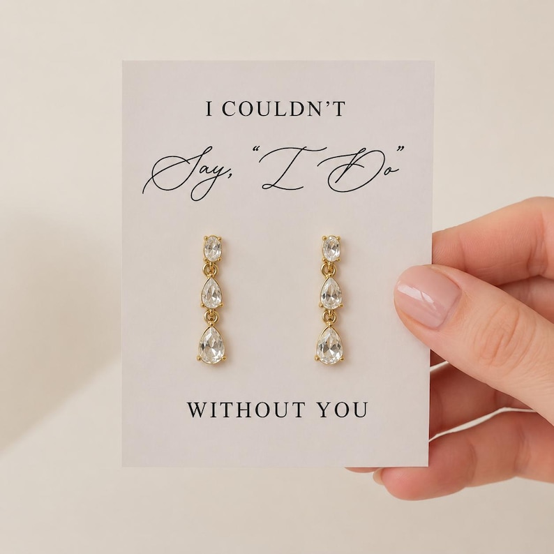 May include: A pair of gold-toned drop earrings featuring three clear, pear-shaped stones on a white display card. The card displays the black text: I COULDN'T Say, "I Do" WITHOUT YOU. The earrings are arranged vertically with a minimalist metal setting.