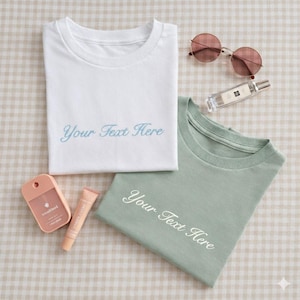 May include: A white t-shirt with blue cursive text reading Your Text Here, a sage green t-shirt with white cursive text reading Your Text Here, round rose-tinted sunglasses, a clear glass perfume bottle, a peach hand sanitizer, and a peach lip balm.