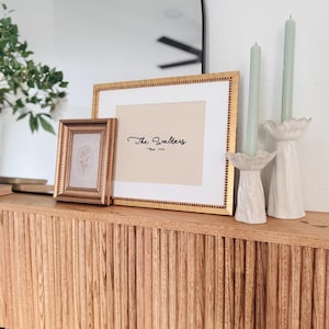 May include: A large rectangular gold frame with a white mat containing the text The Walters Est. 1986, a smaller square gold frame with a floral print, and two white ceramic candle holders with sage green taper candles on a wooden surface.