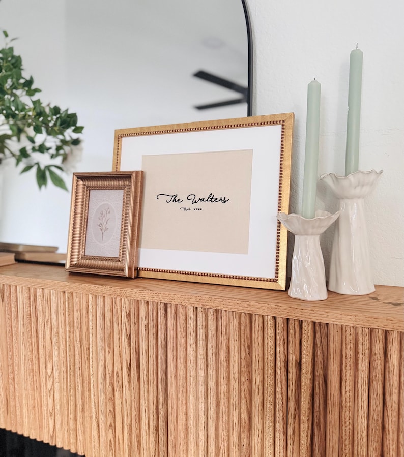 May include: A large rectangular gold frame with a white mat containing the text The Walters Est. 1986, a smaller square gold frame with a floral print, and two white ceramic candle holders with sage green taper candles on a wooden surface.
