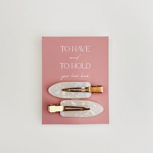 May include: Two pearlescent white hair clips with gold-colored metal clasps are displayed on a pink card. The card has the text "TO HAVE and TO HOLD your hair back" in a handwritten style font.