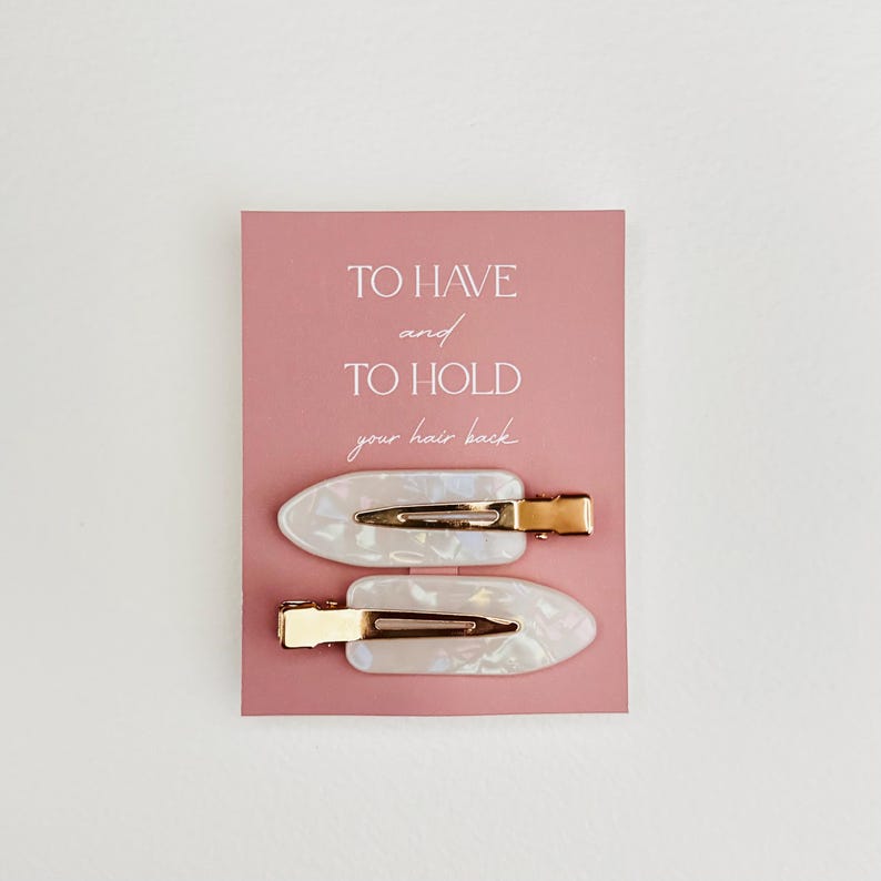 May include: Two pearlescent white hair clips with gold-colored metal clasps are displayed on a pink card. The card has the text "TO HAVE and TO HOLD your hair back" in a handwritten style font.