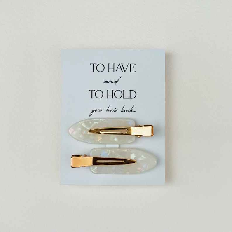 May include: Two pearlescent white hair clips with gold-colored metal clasps are displayed on a light blue card. The card has the text "TO HAVE and TO HOLD your hair back" in black script.