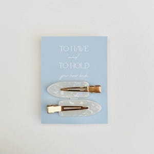 May include: Two white oval hair clips with gold-colored metal clasps are displayed on a light blue card. The card has the text "TO HAVE and TO HOLD your hair back" in a handwritten style font.