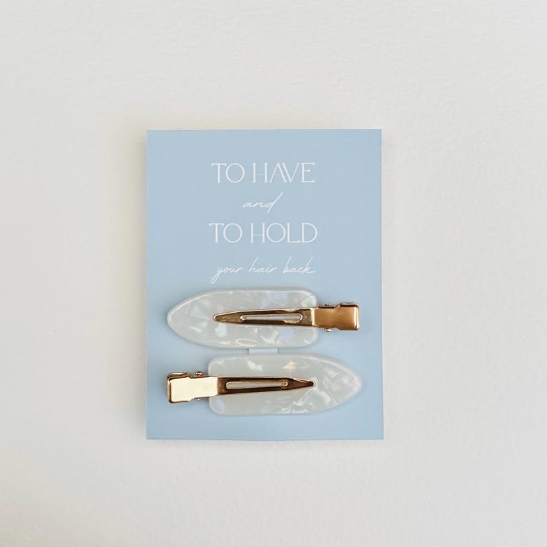May include: Two white oval hair clips with gold-colored metal clasps are displayed on a light blue card. The card has the text "TO HAVE and TO HOLD your hair back" in a handwritten style font.