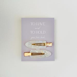 May include: Two pearlescent white hair clips with gold-colored metal clasps are displayed on a light purple card. The card has the text "TO HAVE and TO HOLD your hair back" in a simple font.