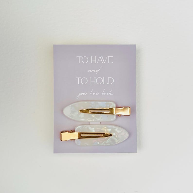 May include: Two pearlescent white hair clips with gold-colored metal clasps are displayed on a light purple card. The card has the text "TO HAVE and TO HOLD your hair back" in a simple font.