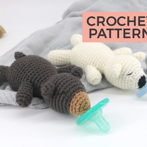 May include: Two crocheted stuffed animals, a brown bear and a white polar bear, are lying on a grey blanket. Each animal has a pacifier in its mouth. The text "CROCHET PATTERN" is displayed in pink on a white background.